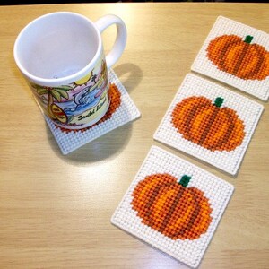 May include: Four white square coasters with orange and green embroidered pumpkins. The coasters are on a light brown wooden surface. A white mug with a beach scene is sitting on one of the coasters.