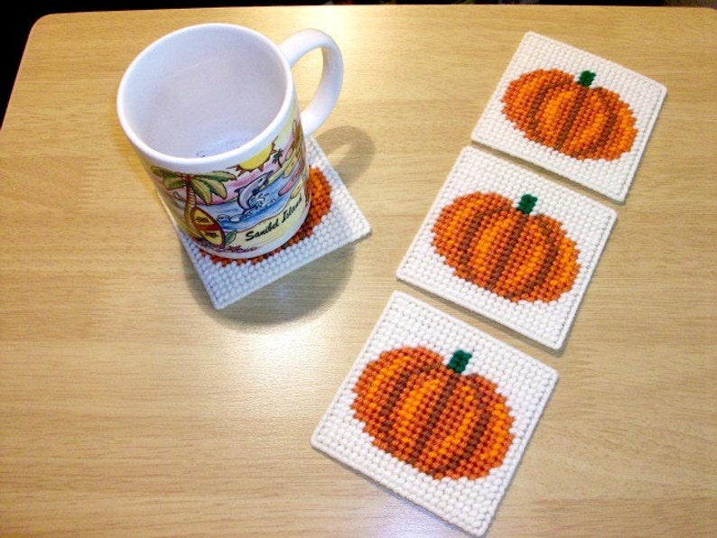 May include: Four white square coasters with orange and green embroidered pumpkins. The coasters are on a light brown wooden surface. A white mug with a beach scene is sitting on one of the coasters.