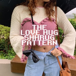 Puede incluir: Una estola de ganchillo rosa y blanca con un diseño en forma de corazón. La estola tiene un borde con volantes y el texto "THE LOVE HUG SHRUG PATTERN" está superpuesto en la imagen.