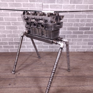 May include: A glass-top table with a unique industrial design. The table base is made from a repurposed engine block and features four metal legs.