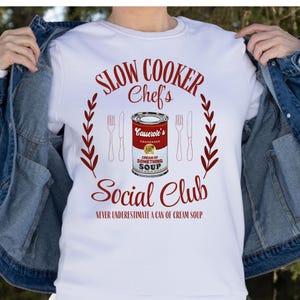 Peut inclure: T-shirt blanc avec du texte rouge qui dit "Slow Cooker Chef's Social Club" et "Ne sous-estimez jamais une boîte de soupe à la crème". Le design présente également une boîte rouge et blanche de crème de quelque chose de soupe condensée avec une couronne rouge et blanche de chaque côté.