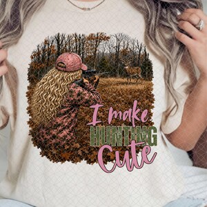May include: A cream-colored t-shirt featuring a graphic of a person with long blonde hair wearing a pink cap and camouflage clothing, aiming a rifle at a deer in a forest. The text reads I make HUNTING CUTE in pink and olive green lettering.