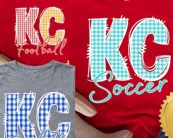 May include: Three t-shirts with the letters "KC" in a gingham pattern. One red shirt has yellow gingham "KC" with "Football" text. Another red shirt has blue gingham "KC" with "Soccer" text. A gray shirt has blue gingham "KC" with "Baseball" text.