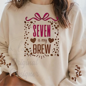 Könnte beinhalten: Beiges Sweatshirt mit dem Text "SEVEN is my BREW" in Pink und Braun. Das Design umfasst Kaffeebohnen, eine rosa Schleife und zwei herzförmige Kaffeebohnen-Designs. Ein lässiges Oberteil zum Thema Kaffee.
