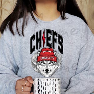 May include: Gray sweatshirt featuring the word "CHIEFS" in black block letters with a red lightning bolt. Below the word is a wolf wearing a red beanie with "KANSAS CITY" on it. The sweatshirt is a comfortable, casual style.