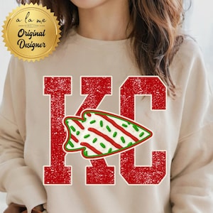 May include: A beige sweatshirt featuring a distressed red "KC" graphic with a green and white arrowhead design. The sweatshirt also has a gold seal that says "a la me Original Designer".