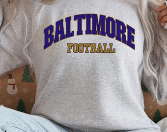 May include: A light gray sweatshirt with the word "BALTIMORE" arched across the chest in dark blue letters with a gold outline. Below the word "BALTIMORE" is the word "FOOTBALL" in gold letters.