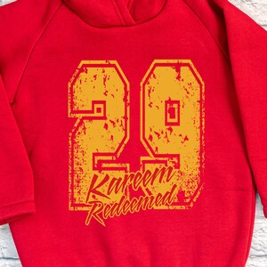 May include: Red hoodie with a distressed yellow number 29 and the text "Kareem Redeemed" printed on the front.