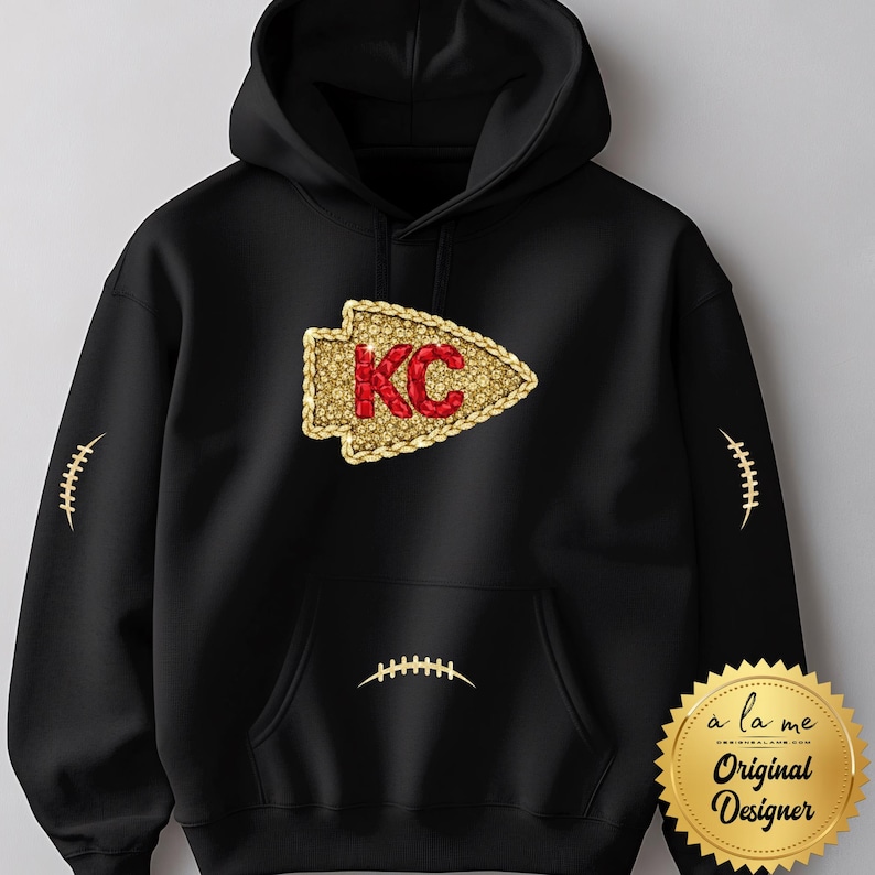 May include: Black hooded sweatshirt featuring a gold and red arrowhead design with the letters "KC". The sweatshirt has football stitching details on the sleeves and pocket. A gold seal with the text "a la me Original Designer" is on the bottom right.