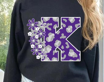 以下が含まれることがあります： 紫とグレーのレオパード柄のKの文字と、紫と白の花の集まりがプリントされた黒いスウェットシャツです。