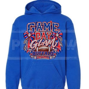 May include: A royal blue hooded sweatshirt featuring a graphic print with the text "GAME DAY GLAM" and "BUFFALO FOOTBALL". The design includes a brown football, red and blue bows with leopard print accents, and sparkling star embellishments.