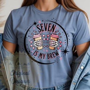 May include: A light blue t-shirt featuring a graphic with two coffee cups held by skeleton hands. The text "SEVEN IS MY BREW" is in pink and white. The design includes stars and geometric shapes.