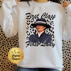 May include: White sweatshirt with a black and white leopard print design. The sweatshirt features a black and white illustration of a woman wearing a black hat and a black suit. The text "First Class First Lady" is printed in black on the sweatshirt.