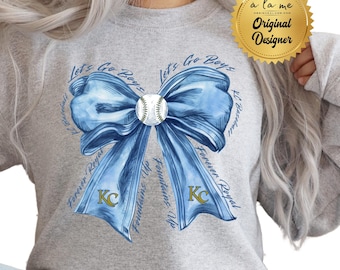 Può includere: Una felpa grigia con un fiocco blu e una palla da baseball al centro. Il fiocco ha le parole "Let's Go Boys" scritte su ogni lato. Le parole "Forever Royal" e "Fountains Up" sono scritte sulle code del fiocco. Le lettere "KC" sono al centro del fiocco.