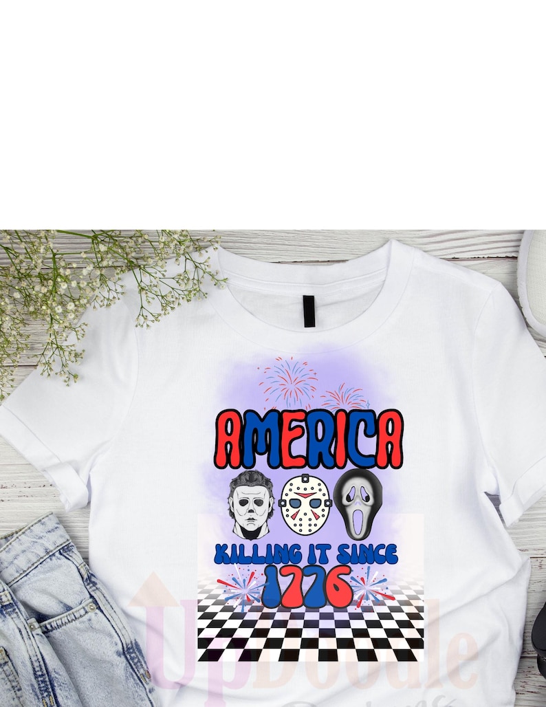 May include: White t-shirt with a graphic design featuring the word "AMERICA" in red and blue, along with images of three horror movie characters. The text "KILLING IT SINCE 1776" is also included.
