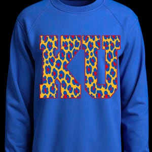 May include: A royal blue sweatshirt with the letters "KU" prominently displayed. The letters feature a yellow leopard print design with red heart accents and a red outline, creating a striking visual.