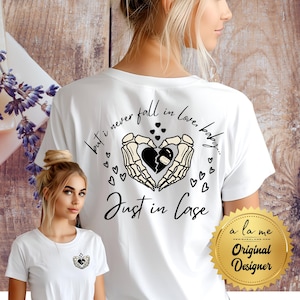 Op de afbeelding: Wit t-shirt met een grafisch ontwerp met een gebroken hart dat wordt vastgehouden door skeletachtige handen, omringd door kleine hartjes. De tekst "but i never fall in love, baby... Just in Case" staat op het shirt.