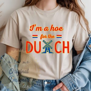 May include: Beige t-shirt with the text "I'm a hoe for the Dutch" in red and blue. The word "Dutch" features a windmill graphic. The shirt also has the text "a la me Original Designer" in a script font.