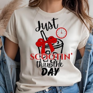 May include: Cream-colored t-shirt with a graphic of a drink with a red bow and a clock. The text on the shirt reads "Just Scootin' thru the DAY" in black and red lettering. The shirt has short sleeves.