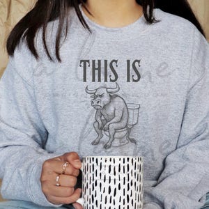 Puede incluir: Una sudadera gris claro con el texto "THIS IS" sobre un gráfico de un toro sentado en un inodoro. La persona de la imagen sostiene una taza blanca con guiones verticales negros.