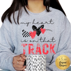 May include: A light gray sweatshirt with the text "my heart is on that TRACK" in black and red. The design includes a black heart, two red hearts, and a checkered flag. A white mug with black dashes is held in the hands.