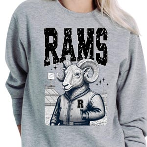May include: Gray sweatshirt featuring a graphic of a ram wearing a letterman jacket with the letter "R". The word "RAMS" is printed above the ram in a distressed, block letter font. The background includes a stadium and stars.