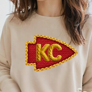 May include: A red arrow-shaped patch with the letters "KC" in gold, outlined with a gold braided border. The patch is on a beige sweatshirt.
