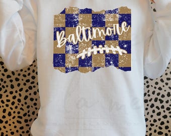 May include: White sweatshirt with a distressed blue and gold checkered design and the word "Baltimore" in white script. A white football stitch design is on top of the checkered design.