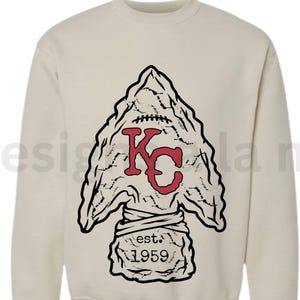 May include: Cream-colored sweatshirt featuring a large arrowhead design. The arrowhead has a football-like texture and a red "KC" logo. Below the arrowhead is the text "est. 1959".
