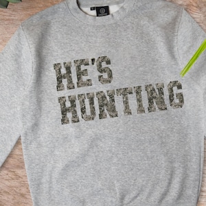 May include: A light gray sweatshirt with the text "HE'S HUNTING" in a camouflage pattern. The sweatshirt has a crew neck and long sleeves. The brand logo is visible on the upper back.