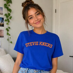 May include: A royal blue short-sleeved t-shirt featuring the text STEVIE KNICKS printed in bold orange capital letters across the chest. The shirt has a classic crew neckline and a straight hemline, constructed from a solid-colored cotton fabric.
