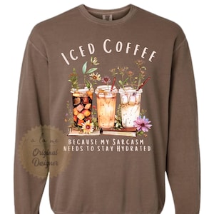 May include: Brown sweatshirt with the words "ICED COFFEE" above a graphic of three iced coffee drinks with flowers and the text "BECAUSE MY SARCASM NEEDS TO STAY HYDRATED". The sweatshirt has a crew neck and long sleeves.
