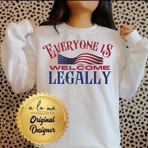 May include: White long-sleeved sweatshirt with the text "EVERYONE IS WELCOME LEGALLY" in red and blue, with an American flag graphic. A gold seal with the text "a la me Original Designer" is at the bottom.