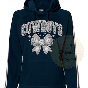 May include: Navy blue hoodie with the word "COWBOYS" in a rhinestone-like font above a large silver bow with rhinestone accents. The hoodie has white stripes down the sleeves and the text "a la me Original Designer".