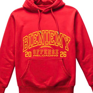 May include: A red hooded sweatshirt with a front pocket and drawstring hood. The sweatshirt features the word "BIENIEMY" in yellow block letters across the chest, with "20 OFFENSE RELOADING 26" below it. The design includes a football graphic.