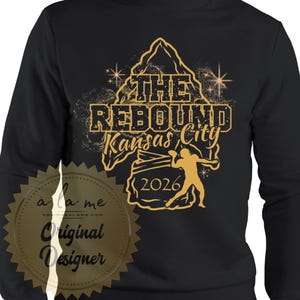 May include: Black sweatshirt with a gold graphic that reads "THE REBOUND Kansas City 2026". The design includes a mountain, stars, and a silhouette of a person throwing a ball. The sweatshirt has a round neckline.
