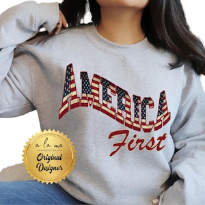 May include: Gray sweatshirt with the text "America First" in red and white. The word "America" is made up of American flag designs. A gold seal with the text "a la me Original Designer" is in the lower left corner.