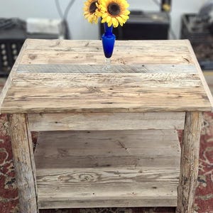 May include: A rustic wooden coffee table with a lower shelf. The table is made from reclaimed wood and has a natural, weathered finish. A blue vase with yellow flowers sits on the top shelf.
