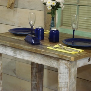May include: A rustic wooden table with a whitewashed finish. The table is set with two blue plates, a yellow napkin, and silverware. There are also two blue salt and pepper shakers on the table.