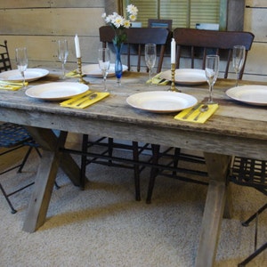 May include: A rustic wooden table with a distressed finish, set for a meal with white plates, yellow napkins, and silverware. The table has a cross-shaped base and is surrounded by black metal chairs.