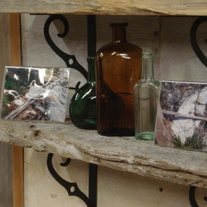 May include: Two rustic wooden shelves with black metal brackets, holding three glass bottles and two framed photographs. The bottles are clear, amber, and green. The photographs are black and white and show nature scenes.