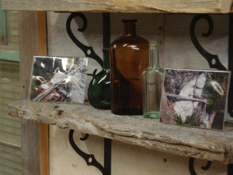 May include: Two rustic wooden shelves with black metal brackets, holding three glass bottles and two framed photographs. The bottles are clear, amber, and green. The photographs are black and white and show nature scenes.
