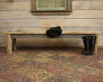May include: A rustic wooden bench with a weathered gray finish sits in front of a wood-paneled wall. The bench has a simple design with two thick legs on each end. A pair of black rubber boots are placed on the floor in front of the bench.