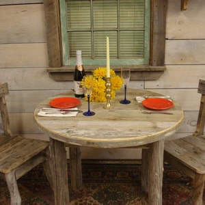 May include: A rustic wooden table with two chairs, set for a romantic dinner. The table has a round top and is made of reclaimed wood. There are two red plates, two glasses, and a bottle of wine on the table. A single candle and a bouquet of yellow flowers complete the setting.