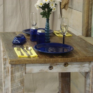 May include: A rustic wooden table with a whitewashed finish, set for a meal with blue and white dishes, silverware, and a yellow napkin. The table has a distressed look with visible wood grain and knots.