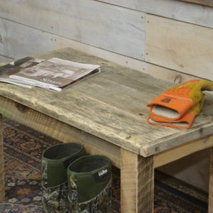 May include: A rustic wooden bench with a weathered gray finish. The bench has a simple design with four legs and a rectangular top. There are two pairs of boots and a pair of work gloves on the floor in front of the bench.