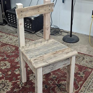 May include: A rustic wooden chair with a light wood finish. The chair is made from reclaimed wood and has a simple design. The chair is sitting on a red and cream patterned rug.
