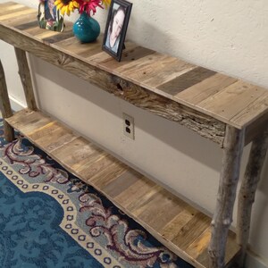May include: A rustic wooden console table with a reclaimed wood top and a lower shelf. The table has two natural wood legs and is decorated with a vase of flowers and a framed photo.
