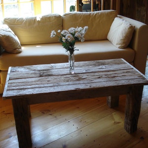 May include: A rustic wooden coffee table with a weathered gray finish. The table has a rectangular top and is supported by four legs. A vase of white flowers sits on the table.