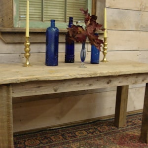 Puede incluir: Una mesa rústica de madera con cuatro patas robustas. La superficie es de color claro y parece estar hecha de madera reciclada. Dos candelabros de latón con velas encendidas, botellas de vidrio azul y un jarrón con hojas rojas están sobre la mesa.